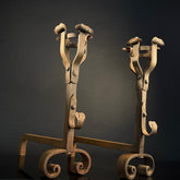 A pair of vintage gothic style hand-beated wrought iron fire dogs with curved legs and a cup-shaped top, showcasing signs of age and use.